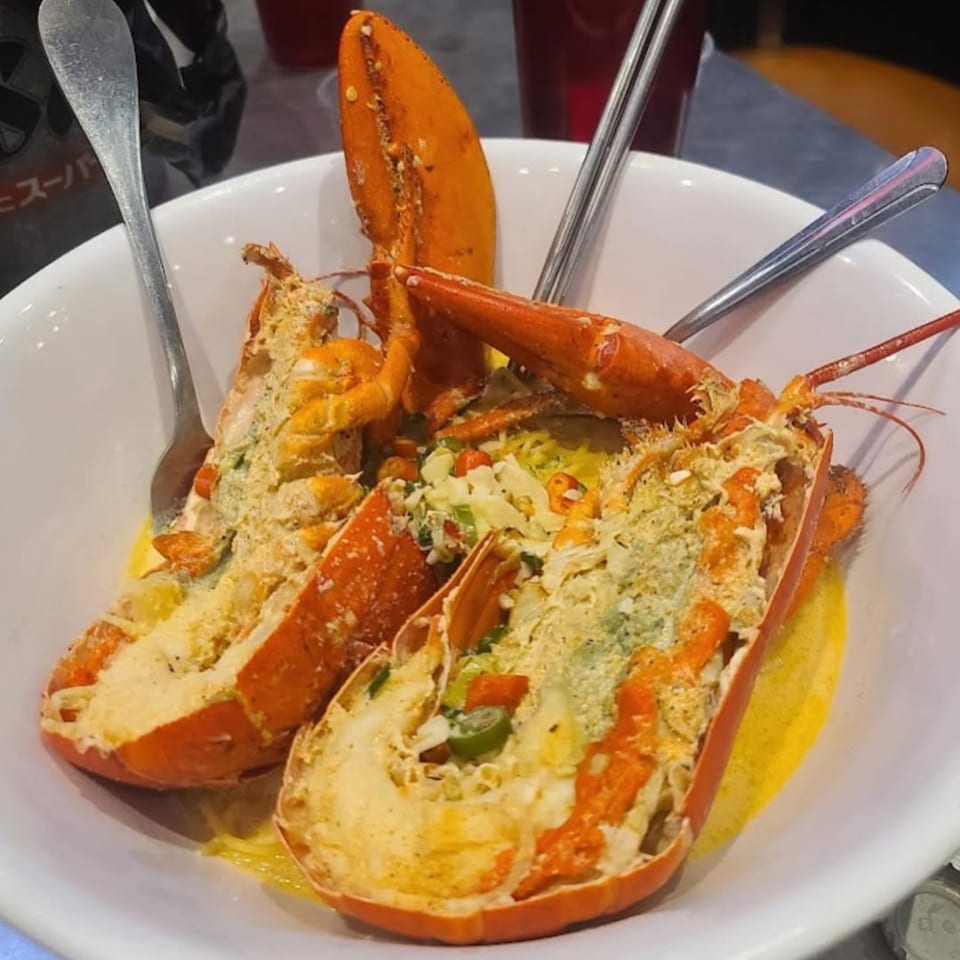 Two halves of baked lobster, topped with cheese and vegetables, served in a white bowl.