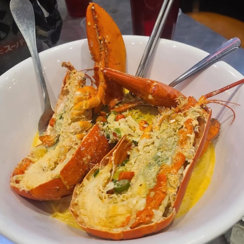 Two halves of baked lobster, topped with cheese and vegetables, served in a white bowl.
